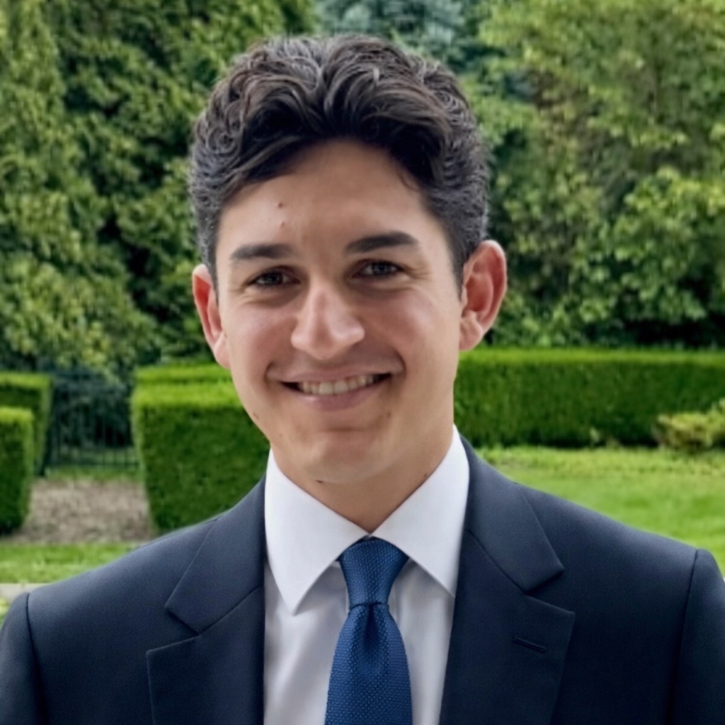 Noah Ahdoot in a navy suit and blue tie, outdoors in a garden